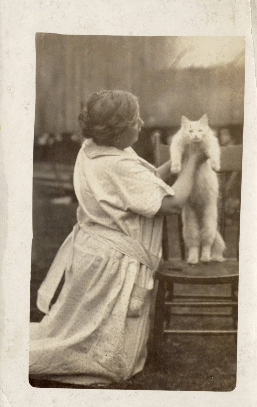 Black and white photo postcard of a woman in a light colored dress. She is sat on her knees and is holding a light colored cat above a chair.